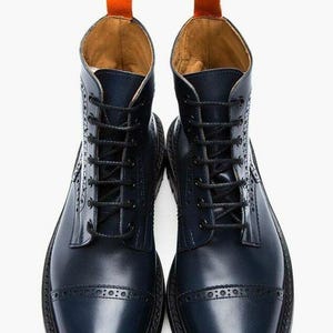 May include: A pair of dark navy blue leather boots featuring a cap toe with decorative brogue perforations, black laces, and a bright orange pull tab at the top of the ankle collar. The boots have a smooth finish and a sturdy black rubber sole.