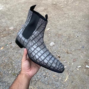 Peut inclure: Une bottine grise avec un motif texturé en peau de crocodile, un panneau latéral élastique noir et deux tirettes noires sur le dessus. La botte présente un bout pointu, un petit talon noir et une silhouette élégante avec des coutures visibles.