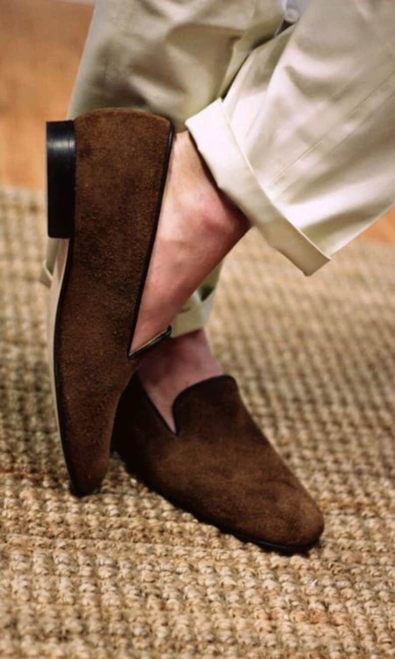 May include: Brown suede slip-on shoes with a black sole and trim. The shoes are worn with beige pants. The shoes have a classic, minimalist design, suitable for casual or semi-formal occasions.