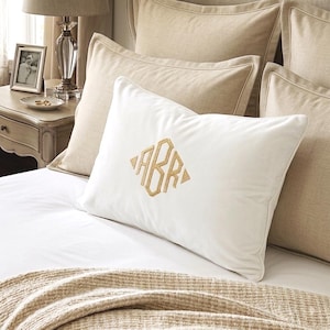 May include: A rectangular white pillow featuring a centred gold embroidered monogram with the letters ABR arranged in a diamond shape. The pillow has a smooth texture and is positioned among several beige pillows on a white bed sheet.