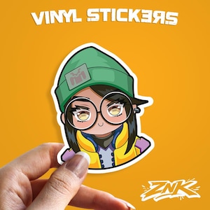 Könnte beinhalten: Ein Vinyl-Aufkleber mit einer Cartoon-Figur mit Brille, grüner Mütze und gelber Jacke. Der Aufkleber hat den Text "VINYL STICKERS" oben und das Logo "ZNK" unten. Der Aufkleber wird von einer Hand vor einem orangefarbenen Hintergrund gehalten.