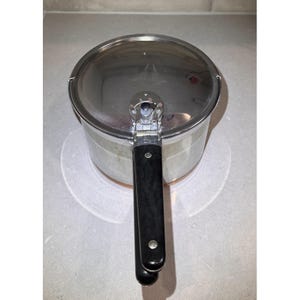 May include: A stainless steel pressure cooker with a polished silver finish and a long, black handle. The lid features a central locking mechanism with a circular dial and metallic accents. The pot has a cylindrical body and a flat, reflective lid.