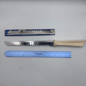 May include: A serrated knife with a cream-colored handle and a metallic blade, positioned next to a blue Westcott ruler measuring 30 centimeters or approximately 12 inches in length, alongside a rectangular box with blue and white graphics.
