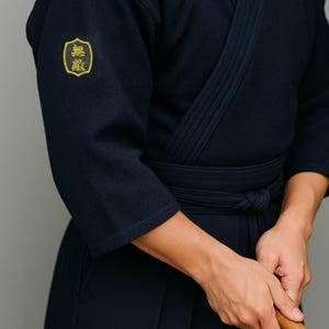 Könnte beinhalten: Marineblauer Kendo-Anzug mit gelbem Emblem auf dem linken Ärmel. Der Anzug hat ein Wickeloberteil und einen passenden Gürtel. Die Person hält ein hölzernes Trainingsschwert.