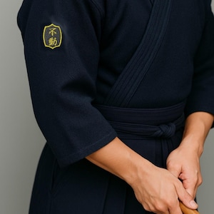 May include: Navy blue Kendo uniform with a yellow embroidered patch on the left sleeve. The uniform features a wrap-style top and a matching belt. The person is holding a wooden sword.