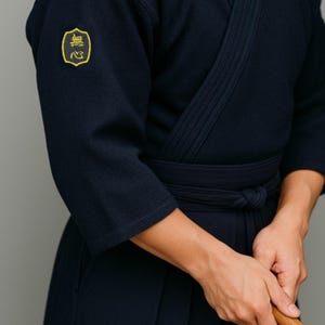 Pode incluir: Uniforme de Kendo azul marinho com um emblema amarelo na manga esquerda. O uniforme apresenta uma parte superior envolvente e um cinto a condizer. A pessoa está segurando um bokken de madeira, uma espada de prática usada no Kendo.