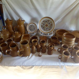 May include: A large collection of brown stoneware dinnerware including multiple mugs, pitchers, lidded pots, bowls, and plates. Several pieces feature a central circular pattern with intricate floral and geometric motifs in shades of brown, cream, and dark orange.