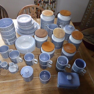 May include: A comprehensive blue and white ceramic tableware set, featuring mugs, plates, bowls, and lidded canisters. The items are displayed on a wooden table, presenting a coordinated design with a decorative pattern.