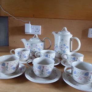 May include: A ceramic tea set featuring a teapot, a coffee pot, and four teacups with matching saucers. Each piece is off-white and decorated with a delicate floral pattern in shades of blue, pink, and green, including small flowers and leaves.
