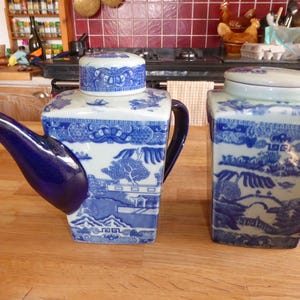 May include: A square ceramic teapot and a matching square canister, both featuring a blue and white willow pattern. The teapot has a dark blue spout and handle, while the canister has a flat lid. Both pieces are decorated with detailed landscape scenes.