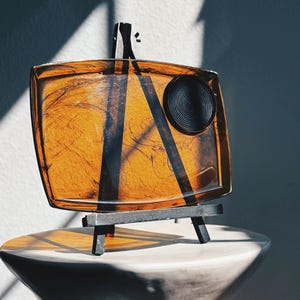 Puede incluir: Una bandeja decorativa cuadrada de color ámbar con detalles negros y un detalle circular, exhibida sobre un pequeño caballete negro. La bandeja se encuentra sobre un pedestal blanco en forma de reloj de arena, creando una exhibición moderna y artística.