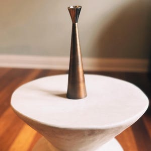 May include: A bronze-colored metal candlestick holder with a conical shape and a silver-colored top. The candlestick is placed on a white, round-topped side table with a flared base. The background features a wooden floor and a neutral-colored wall.