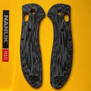 May include: Two black and grey textured knife handle scales featuring a wood grain pattern. Each scale has a large circular hole at the top, a smaller oval hole, and several smaller circular screw holes along the edges and base.