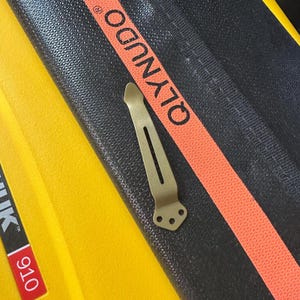 May include: A matte olive green metal pocket clip featuring a tapered shape with a central vertical slot and a base with three small circular screw holes. The clip is positioned on a black textured surface with the orange text QLYNUDO printed.