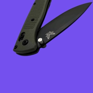 May include: A folding pocket knife with an olive green textured handle and a matte black blade. The blade features a silver thumb stud and the Benchmade logo printed in white near the base. The handle has visible black screws and a pivot pin.