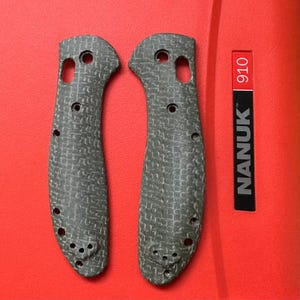 May include: Two dark gray knife handle scales featuring a textured, cross-hatched pattern. Each scale has multiple pre-drilled holes for assembly and a curved shape. Next to the scales is a black label with the white text NANUK 910.