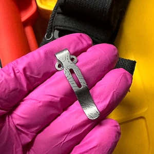 May include: A silver-colored metal pocket clip featuring a dark, etched topographic map pattern. The clip has a curved top section with two circular mounting holes and a long, narrow vertical slot running down the center of the main body.