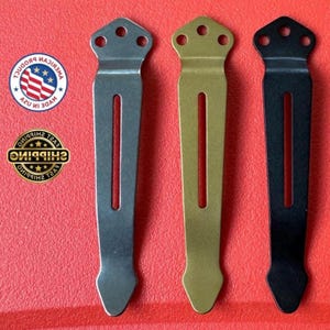 May include: Three metal pocket clips in silver, gold, and black finishes. Each clip features a flat top section with three circular holes for mounting and a long, narrow vertical slot running down the center of the body, ending in a pointed tip.