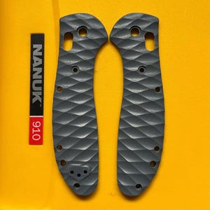 May include: Two dark gray knife handle scales featuring a textured diamond pattern. Each scale includes pre-drilled holes for assembly and a pocket clip mounting point on one side. A black and red label with the text NANUK 910 is visible nearby.