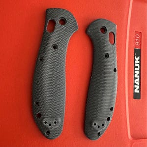 May include: Two black textured knife handle scales with a cross-hatched pattern, featuring pre-drilled holes for assembly and hardware. The scales are positioned side-by-side against a bright red surface with the text NANUK 910 visible.