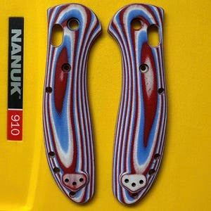 May include: Two knife handle scales featuring a layered, striped pattern in red, white, and blue. Each scale has a curved, ergonomic shape with multiple pre-drilled circular holes for assembly, showcasing a distinct, repeating color-banded design.