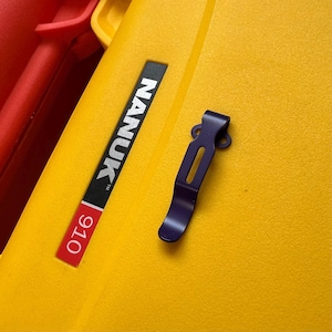 May include: A dark blue metal pocket clip with a central slot and two circular mounting holes at the top, positioned on a textured yellow surface next to a black and red label that reads NANUK 910.
