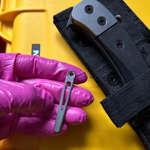 May include: A small, grey, elongated metal clip with a circular hole at the top and a central vertical cutout. The clip is held against a black knife handle secured in a black fabric sheath, positioned over a bright yellow hard-shell case.
