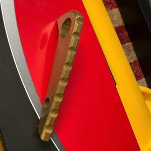 May include: A metallic, brass-colored tool featuring a textured, notched edge along one side and a circular hole at the top. The object has a flat, elongated body with a matte finish and a secondary smaller hole located near the bottom section.