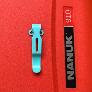 May include: A light blue metal pocket clip featuring a central vertical slot and two circular mounting holes at the top. The clip is positioned next to a red surface with a black and white label that reads NANUK 910 in vertical orientation.