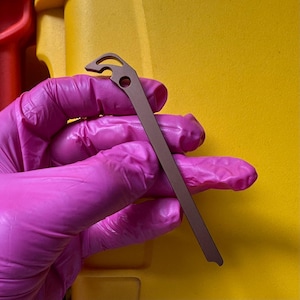 May include: A metallic brown, slender, flat tool with a tapered end and a hook-shaped head featuring a circular cutout. The object is held by a hand wearing a bright pink glove, showcasing the tool's matte finish and its simple, functional design.