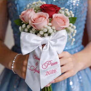 Könnte beinhalten: Ein Blumenstrauß aus rosa und roten Rosen, akzentuiert mit weißem Schleierkraut. Ein großes weißes Satinband ist um die Stiele gebunden und mit den Worten "Olive & Ayden Prom 2026" in rosa Schrift bestickt. Der Strauß wird von einer Person in einem blauen Kleid gehalten.