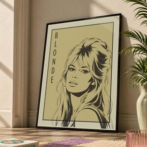 May include: Black framed art print featuring a stylized illustration of a woman with the word "BLONDE" vertically on the left. The artwork is set against a beige background. The print is leaning against a wall on a patterned rug.