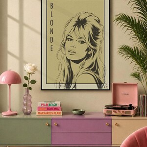 May include: A framed art print featuring a black and white illustration of a woman with the word "BLONDE" vertically printed on the left. The print hangs above a colorful cabinet with books, a record player, and a pink lamp.