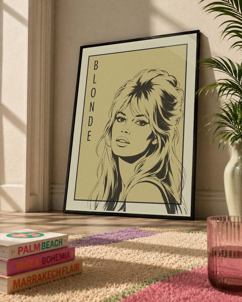May include: Black framed art print featuring a stylized illustration of a woman with the word "BLONDE" vertically on the left. The artwork is set against a beige background. The print is leaning against a wall on a patterned rug.