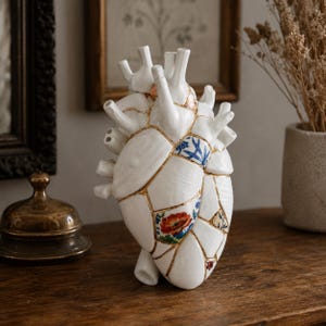 May include: A white ceramic anatomical heart sculpture featuring a kintsugi-style design with gold-filled cracks. Several segments of the heart are decorated with blue and white floral patterns, a red poppy motif, and intricate gold-toned details.