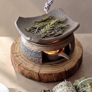 Puede incluir: Un difusor de aceites esenciales de cerámica gris con un plato cuadrado que contiene hierbas secas y lavanda. El difusor se encuentra sobre una base de madera con una vela encendida en su interior. La estética general es rústica y natural.