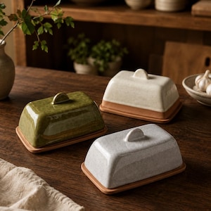 May include: Three ceramic butter dishes in green, white, and gray, each with a lid and a terracotta-colored base. The butter dishes are rectangular with rounded edges and a small handle on top. They are arranged on a dark wooden table.