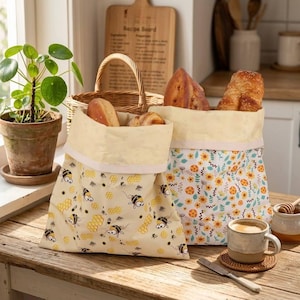 May include: Two fabric bread bags with folded-over tops, filled with loaves of bread. One bag features a bee and honeycomb pattern on a cream background, the other has a floral design. A cup of coffee and a plant are also visible.