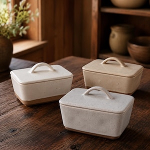 Op de afbeelding: Drie rechthoekige keramische botervlootjes met deksel. De schalen zijn gebroken wit met gespikkelde details en een bruine basis. Elk heeft een gebogen handvat op het deksel. Geplaatst op een houten tafel.