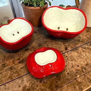 May include: Three ceramic apple-shaped bowls with a glossy red exterior and a white interior. The bowls have a seed detail and a green stem. One bowl is a lid. The bowls are on a wooden surface.