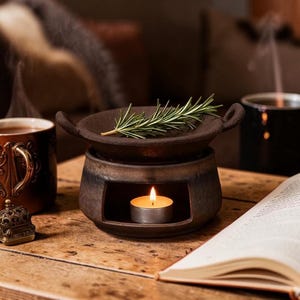 Op de afbeelding: Een donkerbruine keramische etherische olieverspreider met een brandende waxinelichtkaars erin. Een takje rozemarijn rust op de bovenste schaal. Een mok thee en een open boek staan ook op de houten tafel.