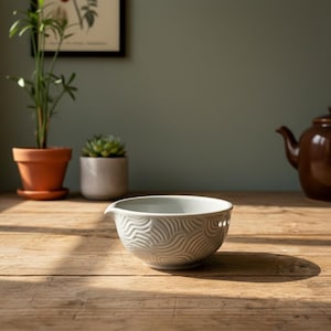 May include: A pale grey ceramic mixing bowl with a spout and a wave pattern. The bowl sits on a wooden surface, with a teapot and plants in the background. The bowl has a small spout for pouring.