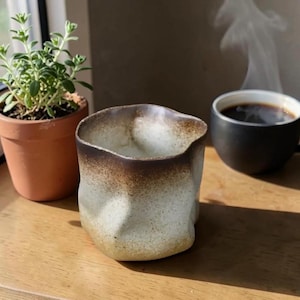 May include: A white ceramic mug with a black handle and the text "But First, Coffee" in a bold, black font. The mug is set against a blurred background, suggesting a cozy setting.