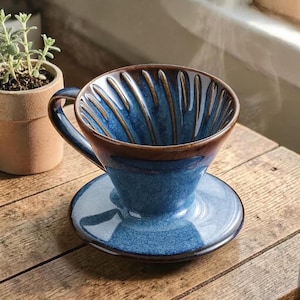 May include: A blue ceramic coffee pour-over set with a matching saucer. The coffee maker has a brown rim and interior, with vertical grooves. The set is on a wooden surface, with a small potted plant in the background.