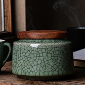 May include: A round, celadon green ceramic jar with a wooden lid. The jar has a crackle glaze pattern. The lid is a dark brown wood. The jar is sitting on a wooden surface. The jar is a decorative item.