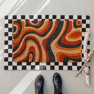 May include: A rectangular doormat with a retro design. The center features swirling patterns in shades of orange, brown, and black. It is framed by a black and white checkered border. Black boots are visible at the bottom.