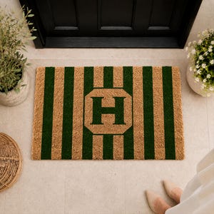 May include: A rectangular doormat featuring a pattern of alternating vertical tan and dark green stripes. In the center, a dark green letter H is printed inside a tan octagonal shape, which is surrounded by a dark green border.