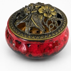 May include: A round ceramic incense burner with a glossy red and black mottled glaze. The vessel features a removable, ornate antique brass-coloured metal lid with a cut-out design of flowers and leaves, revealing a perforated interior base.