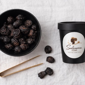 May include: A black bowl of dark, dried fruit sits beside a black container with a white label showing a woman's silhouette and the text "Satourou Gowe". Gold tongs and scattered fruit are also present.
