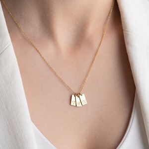 May include: A gold-toned necklace featuring three small, rectangular vertical pendants hanging from a delicate chain. Each pendant is engraved with a single letter, collectively displaying the initials T, R, and M in a classic serif font.